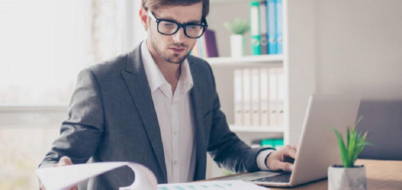 Portrait young handsome businessman with glasses is sitting at the desk in the office workstation and scrutinizing documents